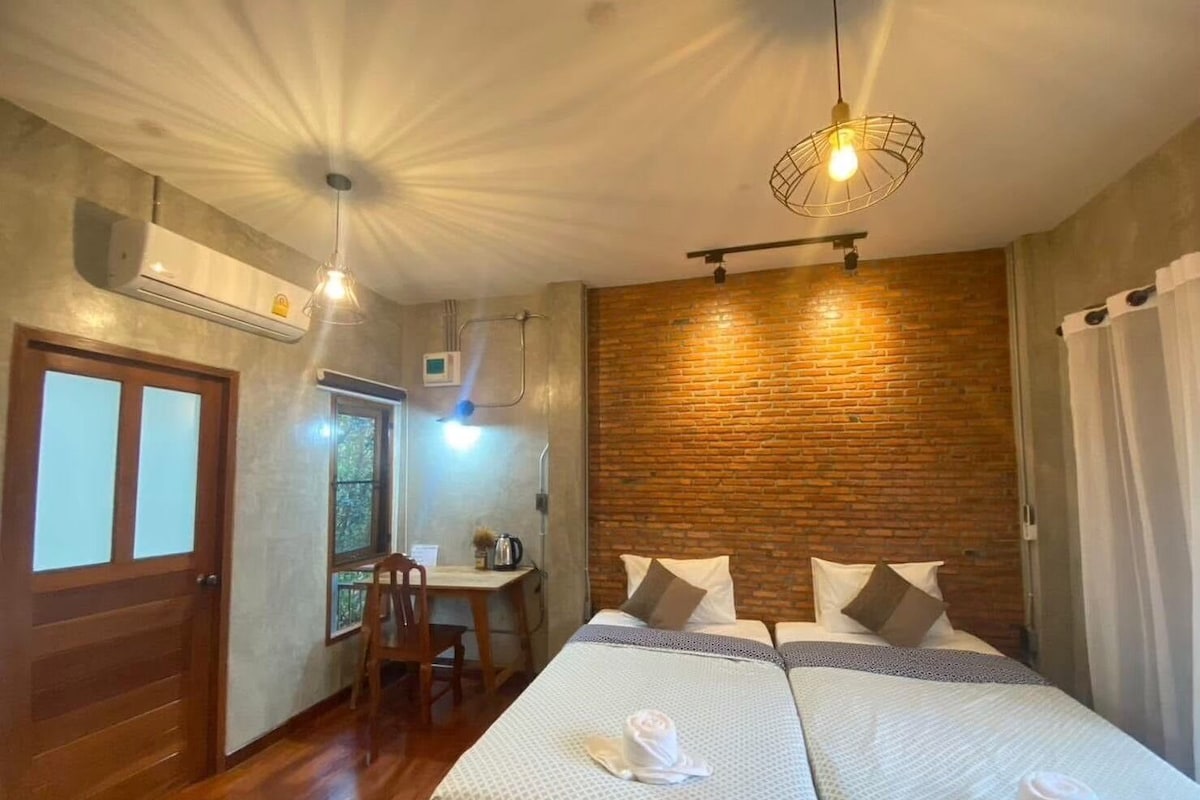 A hotel room with two beds, a wooden floor, a brick wall, and a desk with a chair.