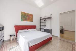 Ein modernes Schlafzimmer mit einem roten Kopfteil, einer weißen Bettdecke, einem kleinen runden Hocker, einem Regal mit Handtüchern und einem farbenfrohen abstrakten Gemälde an der Wand.
