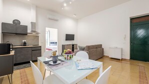 Een moderne keuken met een magnetron, kookplaat en afzuigkap. Er is een eethoek met een tafel, stoelen en een schaal fruit. Een bank en een televisie zijn ook zichtbaar.