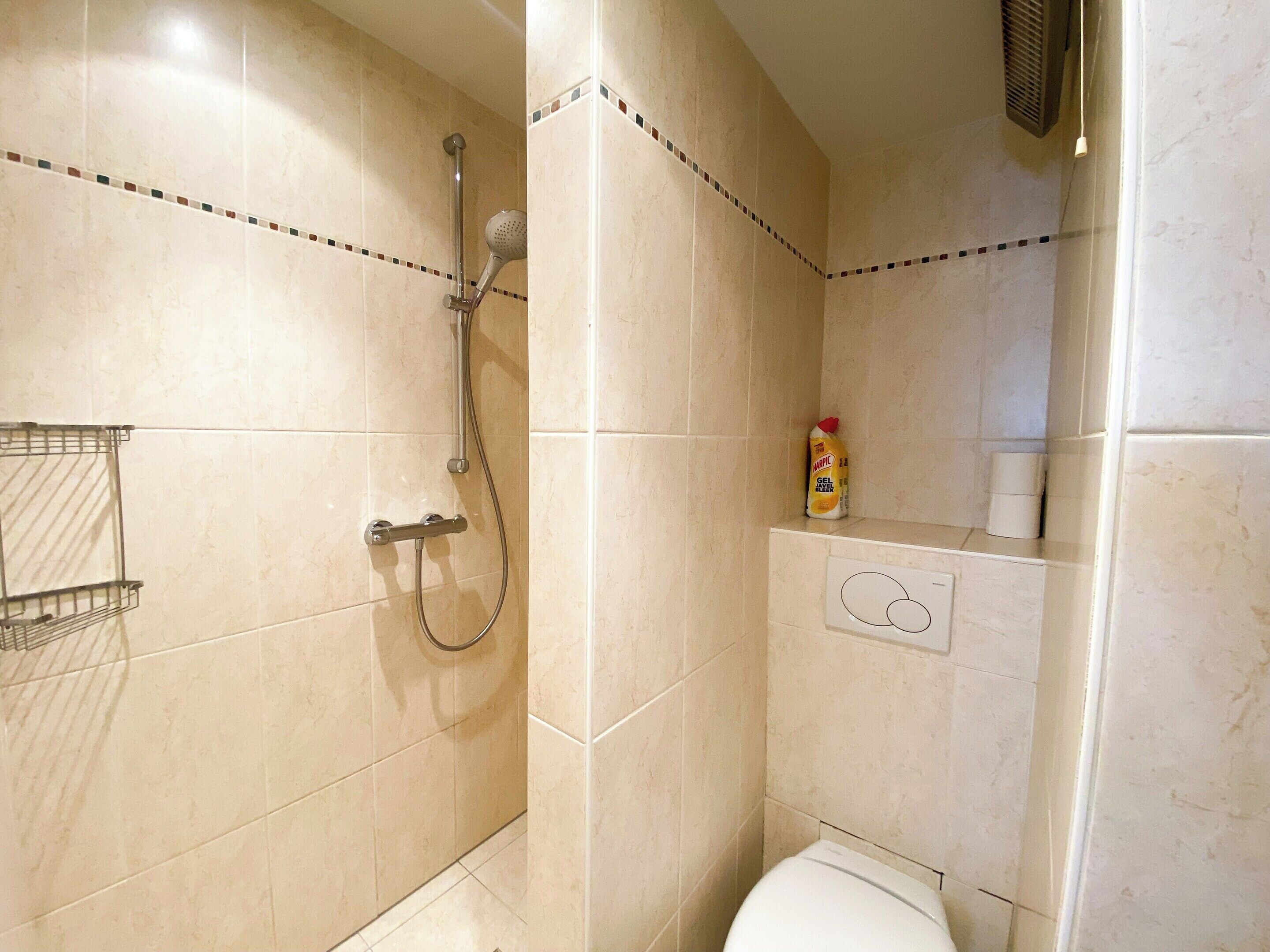 Ein Badezimmer mit beigen Fliesen, einer Dusche mit Duschbrause, einer Toilette und einem Ablagefach für Pflegeprodukte.