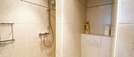 Ein Badezimmer mit beigen Fliesen, einer Dusche mit Duschbrause, einer Toilette und einem Ablagefach für Pflegeprodukte.