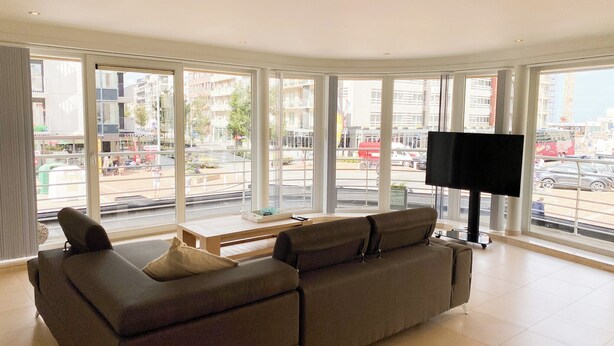 A modern living room with a sofa, a coffee table, and a TV. Large windows offer a view of the city. - Apartment in Nieuwpoort-bad Near Beach (Nieuwpoort)