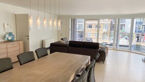 A modern living room with a large dining table, comfortable seating, and large windows offering a view of the city. - Apartment in Nieuwpoort-bad Near Beach (Nieuwpoort)