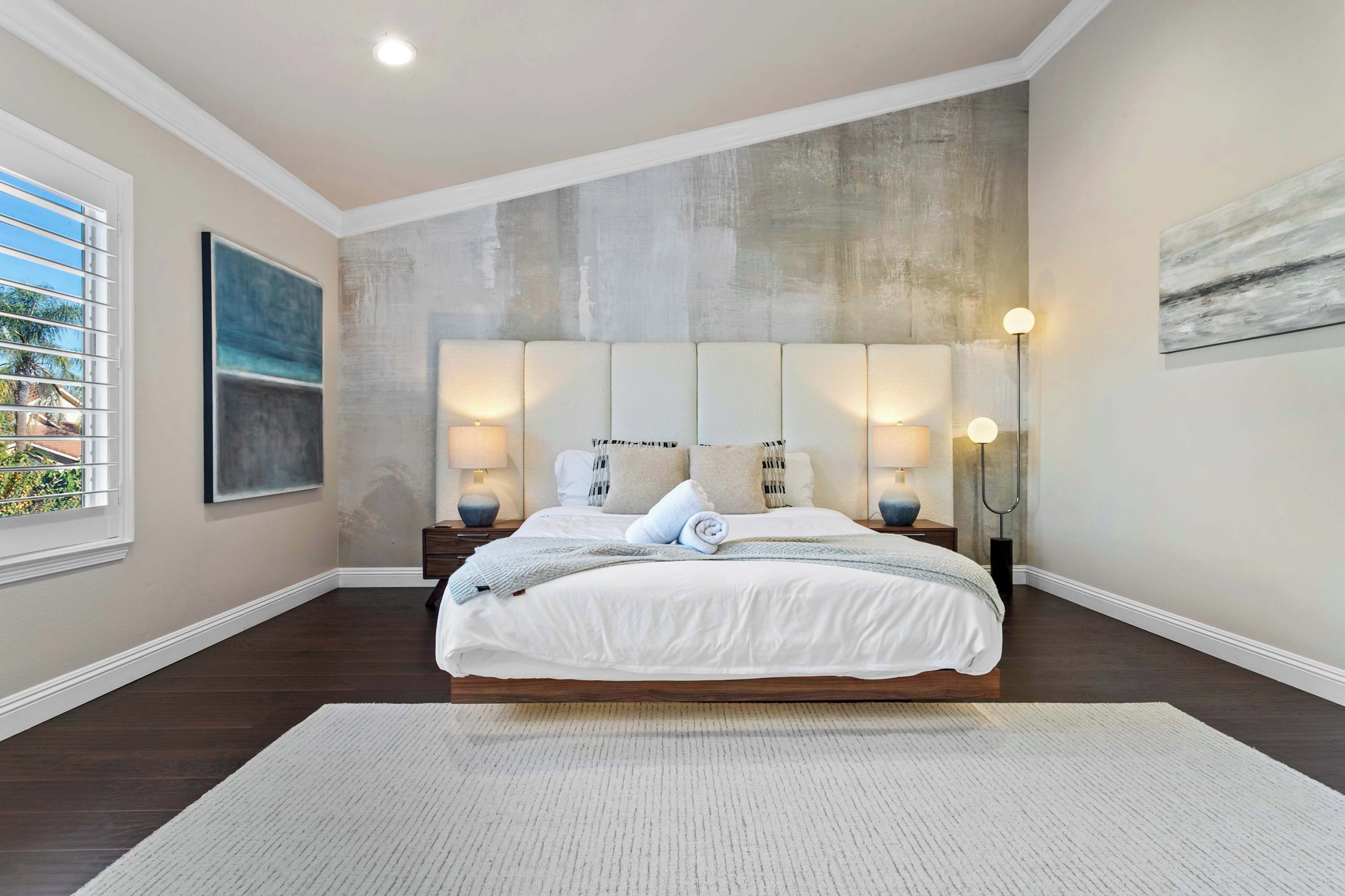 A bedroom with a large bed, two bedside tables, a floor lamp, and a window with blinds.