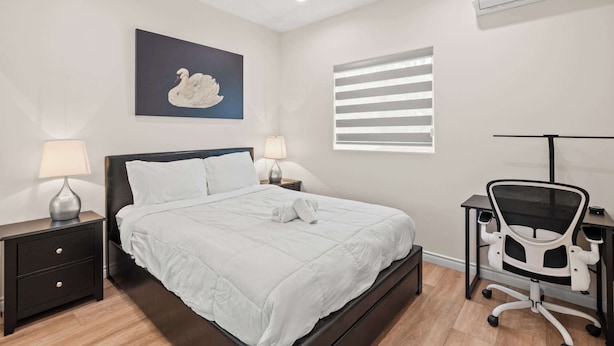 Un dormitorio ordenado con una cama, mesitas de noche, un escritorio con silla, aire acondicionado instalado en la pared y una ventana con persianas.