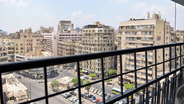 A cityscape viewed from a high-rise building with multiple floors, featuring a mix of residential and commercial buildings, a busy street with cars and pedestrians, and a clear sky. - lora Altahrir hotel (Cairo)