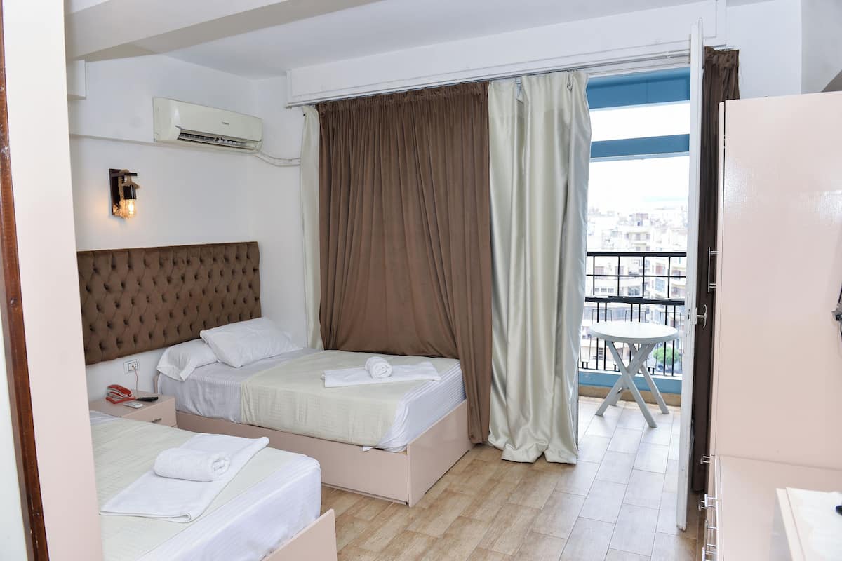 A hotel room with two beds, a large window with curtains, an air conditioning unit, and a balcony with a table.