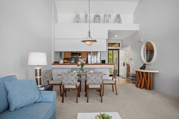A modern living room with a blue sofa, a dining table with chairs, a kitchen area, and a large 'ALOHA' sign above the kitchen.