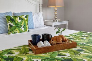 A neatly made bed with a tropical leaf-patterned bedspread, a wooden tray with croissants, a vase with a plant, and a bedside table with a lamp.