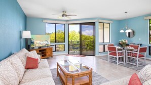 A living room with a sofa, coffee table, and dining area with chairs. There is a ceiling fan and a painting on the wall. - Grand Champions | Ocean View 2 BR, Sleeps 8 | Car Incl. w/ 6+ Nights | GCH ML-1493 by KBM (Wailea)