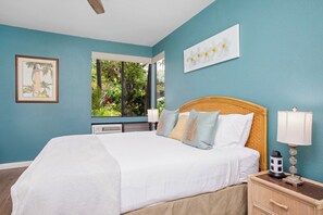 A bedroom with a large bed, a window with a view of greenery, a bedside table with a lamp, and a framed picture on the wall. - Grand Champions | Ocean View 2 BR, Sleeps 8 | Car Incl. w/ 6+ Nights | GCH ML-1493 by KBM (Wailea)