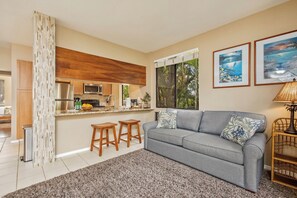 Ein Wohnzimmer mit grauem Sofa, zwei Holzschemeln, einem Couchtisch, einer Lampe und einem durch ein Fenster sichtbaren Küchenbereich.