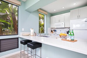A modern kitchen with white cabinets, a large window with a view of greenery, a sink, and two bar stools. - Grand Champions | Ocean View 4 BR, Sleeps 14 | Car Incl. w/ 6+ Nights | GCH ML-1496 by KBM (Wailea)