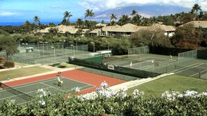 Un complexe de courts de tennis comprenant plusieurs terrains, entouré d’une végétation luxuriante et de palmiers, avec une vue sur une montagne et l’océan en arrière-plan.