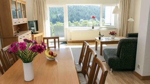 Un coin repas avec une table en bois, des chaises, un vase de fleurs et un saladier de fruits. On y trouve également un téléviseur posé sur un meuble en bois, un canapé et une fenêtre donnant sur des arbres.