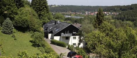 Une maison à deux étages, dotée d’un toit en pente, entourée d’une végétation luxuriante et d’arbres. Deux voitures sont garées dans l’allée.