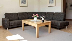 Un salon moderne avec un canapé gris, une table basse en bois ornée d’un vase de fleurs et de deux verres à vin, et des tableaux encadrés accrochés aux murs.