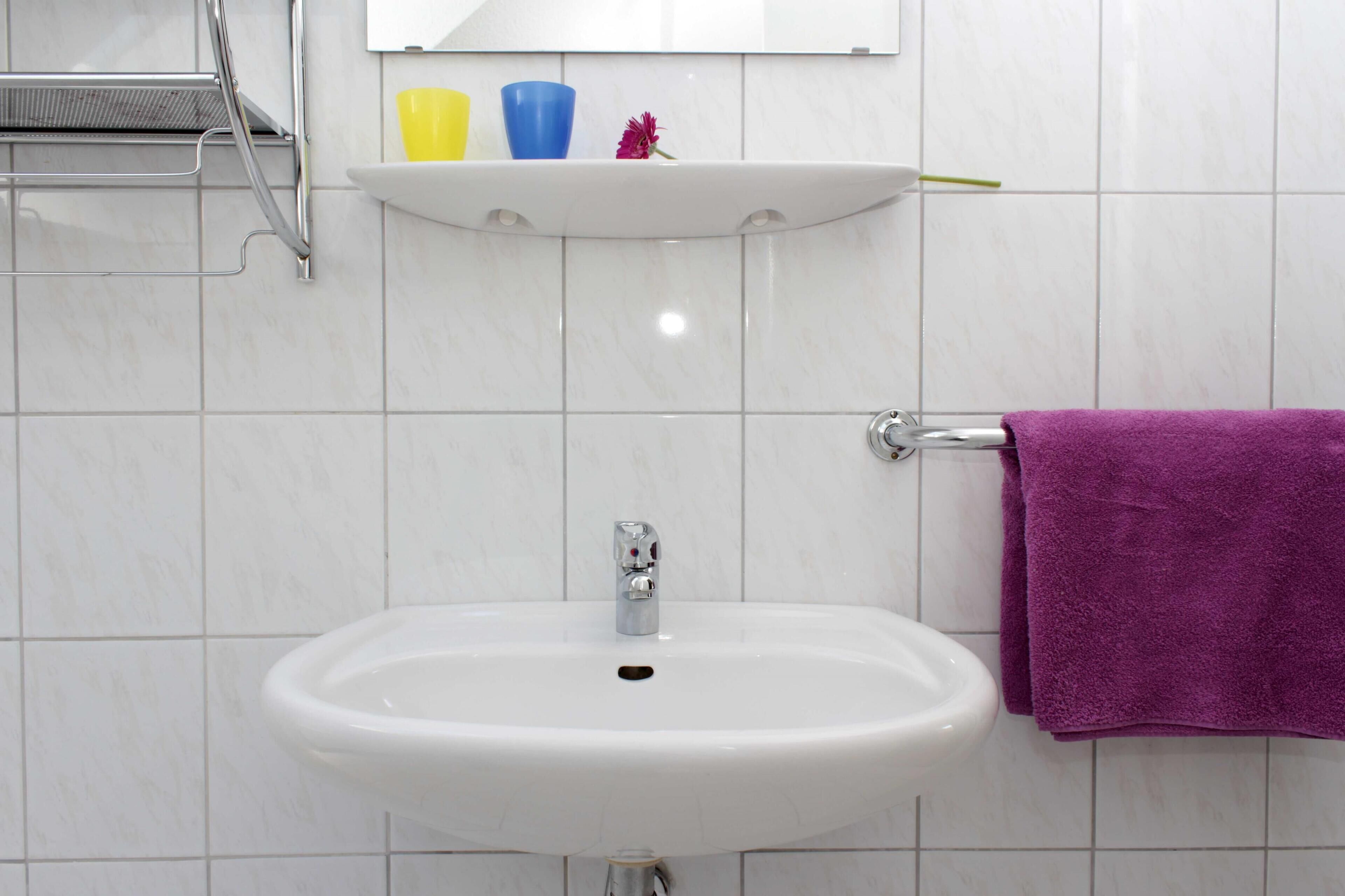 A bathroom with a white sink, a mirror above it, and a shelf with a yellow cup, a blue cup, and a pink flower.