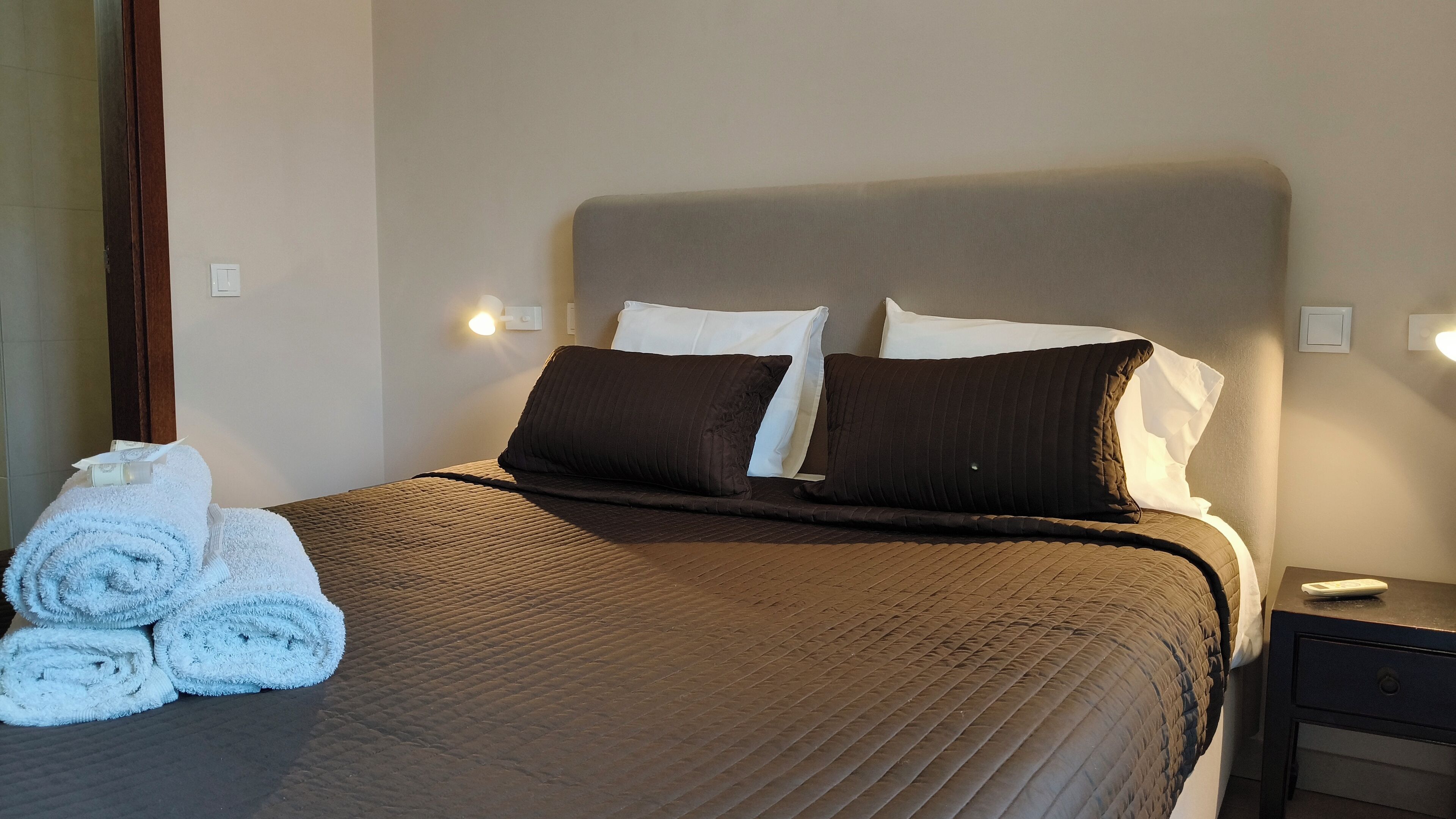 A neatly made bed with a brown bedspread and two pillows, a set of towels, and a bedside table with a remote control.