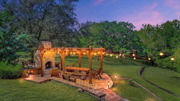 An outdoor patio with a stone fireplace, wooden pergola, and string lights.