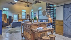 A spacious living area with a wooden island table, ceiling fans, and a large window with blinds.