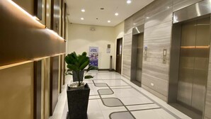 Un hall d’hôtel moderne, avec une plante en pot au centre, un sol carrelé orné d’un motif géométrique et une rangée d’ascenseurs sur le côté droit.