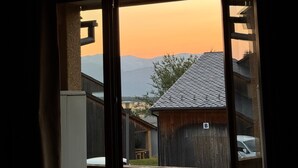 Blick durch eine Glastür auf einen Balkon mit Holzgeländer, ein Gebäude mit Ziegeldach und Berge im Hintergrund.