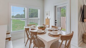 A modern dining area with a white table set for four, wooden chairs, and a view of a green landscape through large windows. - The Modern Cottage near 30a, Golf Cart + Bikes included! (Inlet Beach)
