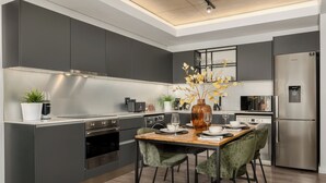 A modern kitchen with dark cabinetry, a stainless steel refrigerator, and a dining area with a wooden table and green chairs. - The Signature | Lush Designer Apartment (Cape Town)