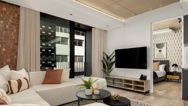 A modern living room with a sofa, a coffee table, and a TV. There is a large window with curtains, a plant, and a bookshelf. - The Signature | Lush Designer Apartment (Cape Town)