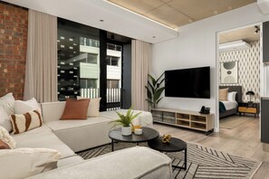 A modern living room with a sofa, a coffee table, and a TV. There is a large window with curtains, a plant, and a bookshelf.