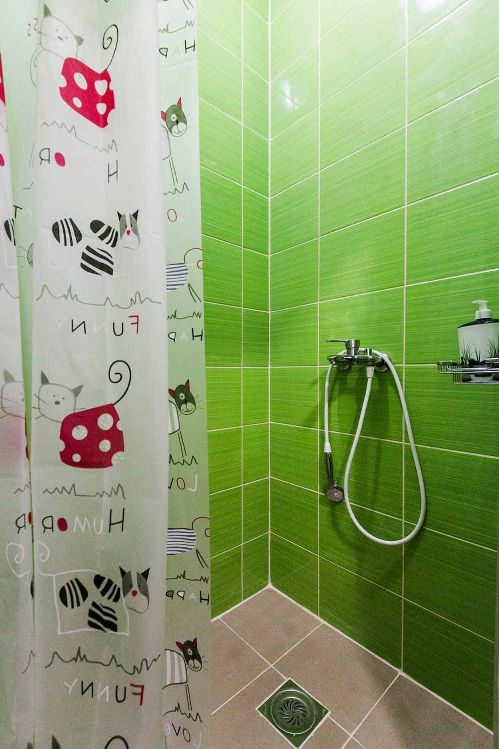 A bathroom with green tiles, a shower with a handheld hose, and a curtain with cat drawings.