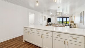 A spacious kitchen with white cabinetry and gold handles, a large island, pendant lighting, and a view of the outdoors.