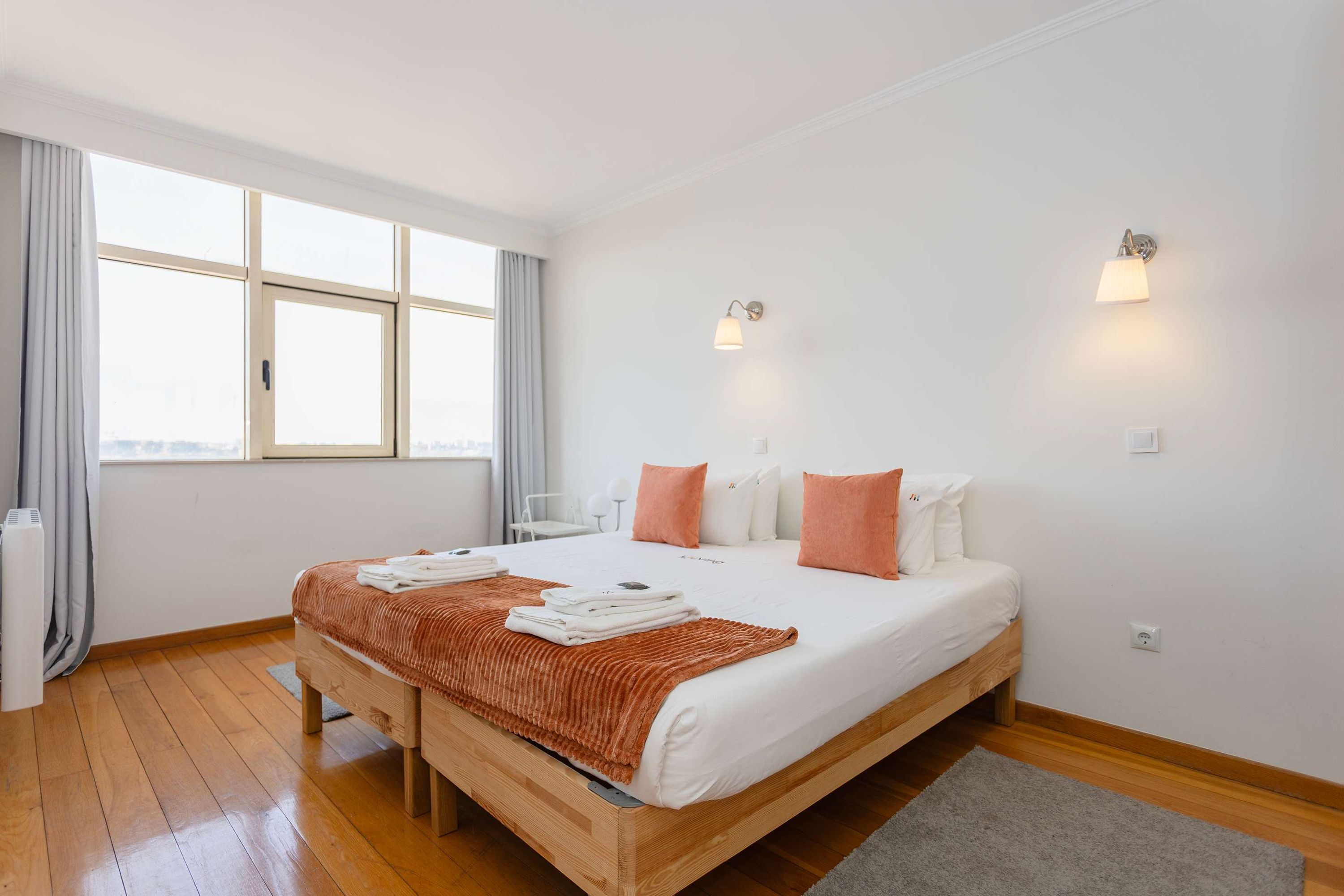 Quarto de hotel moderno com uma cama grande, duas luminárias fixadas na parede, uma janela com cortinas e vista para a cidade.