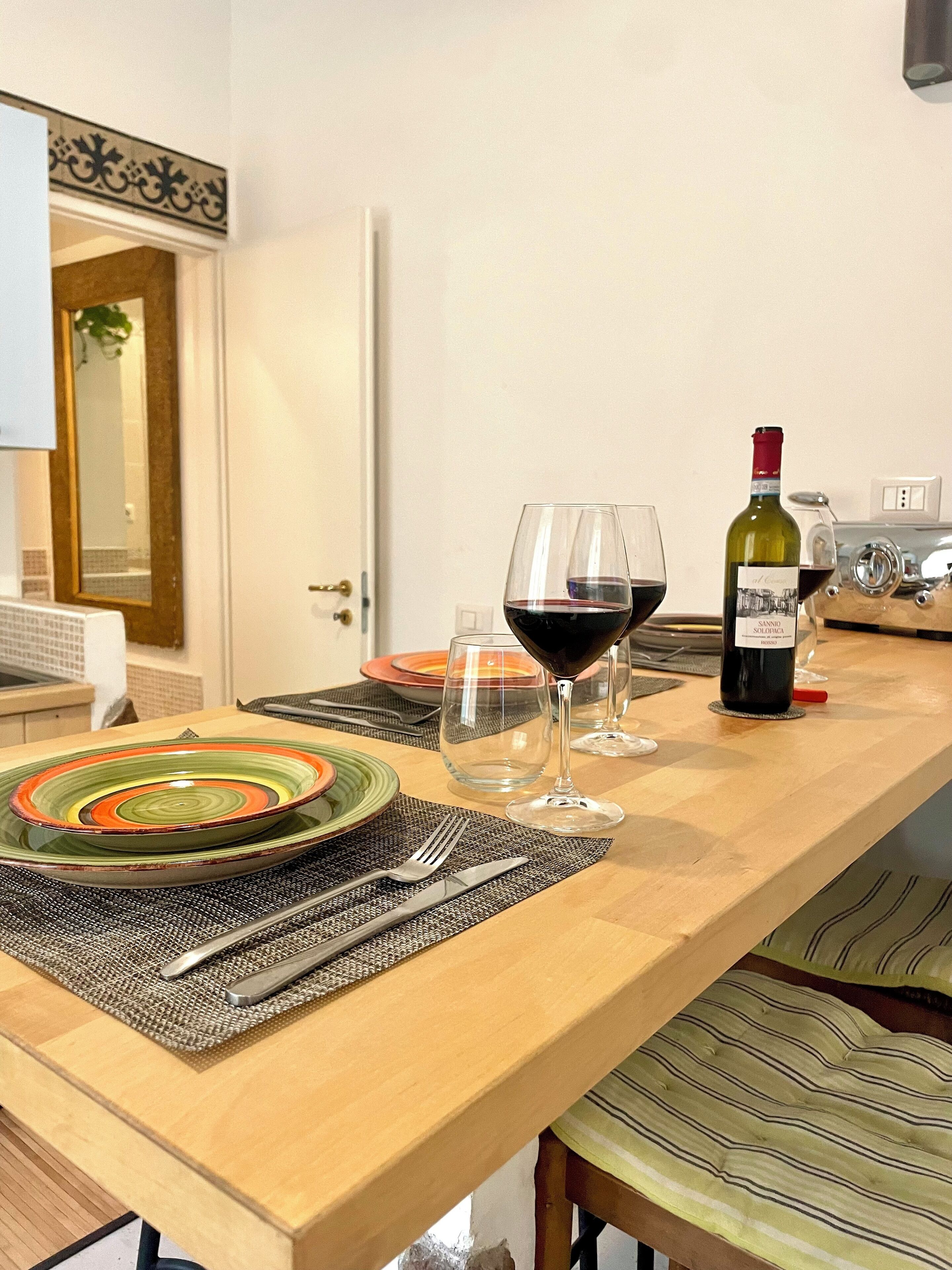 Un comedor con una mesa de madera preparada para dos personas, que incluye una botella de vino, dos copas y un plato colorido.