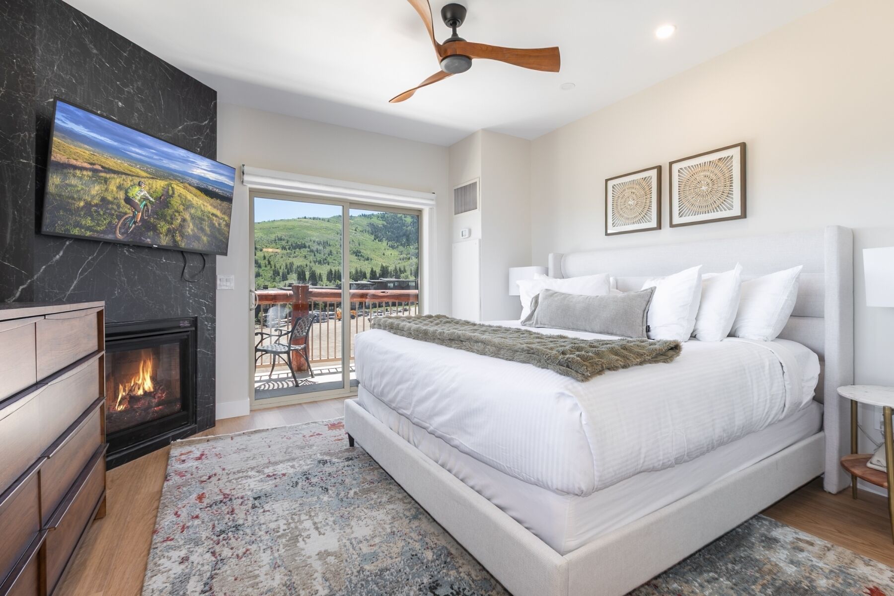 A bedroom with a large bed, a fireplace, a ceiling fan, and a balcony with a mountain view.