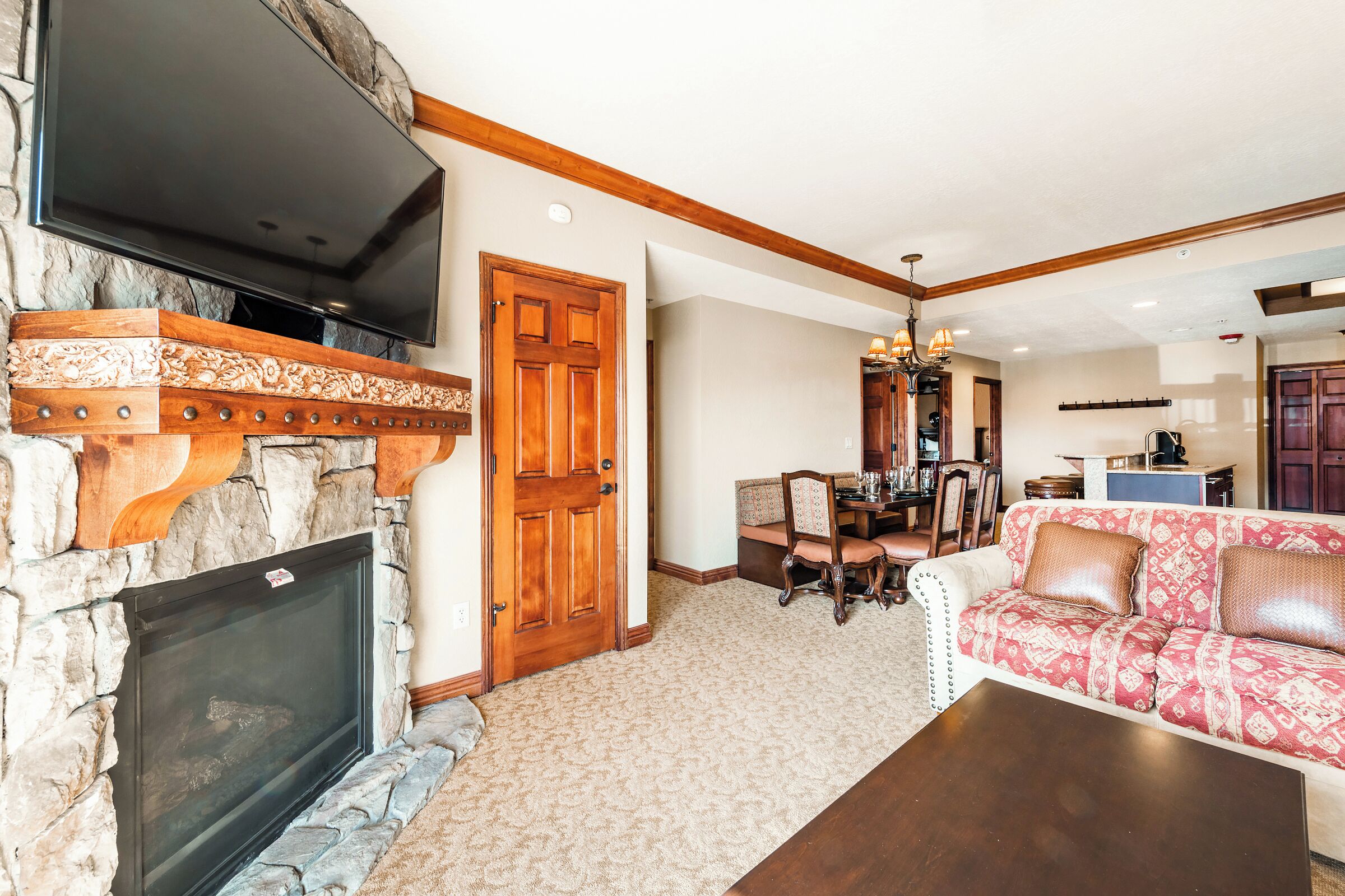 A living room with a stone fireplace, a flat-screen TV mounted above it, a wooden door, a dining area with a chandelier, and a patterned sofa.