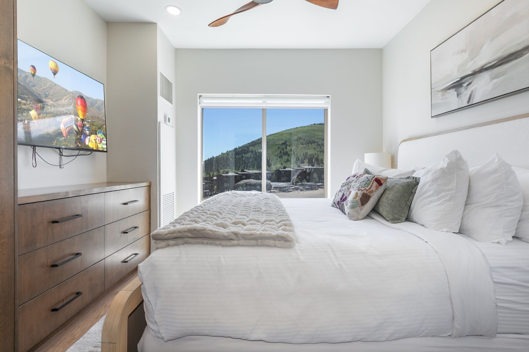 Un dormitorio con cama, televisor, cómoda, ventilador de techo y una ventana grande con vistas a la montaña.