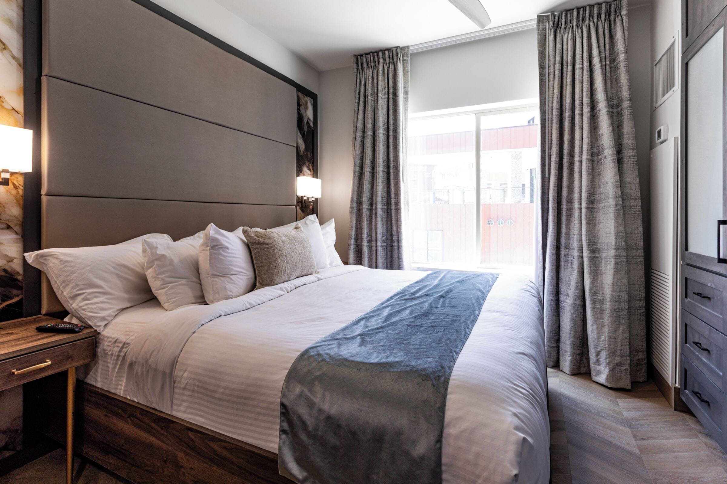 A modern hotel room with a large bed, a nightstand, a dresser, and a window with curtains.