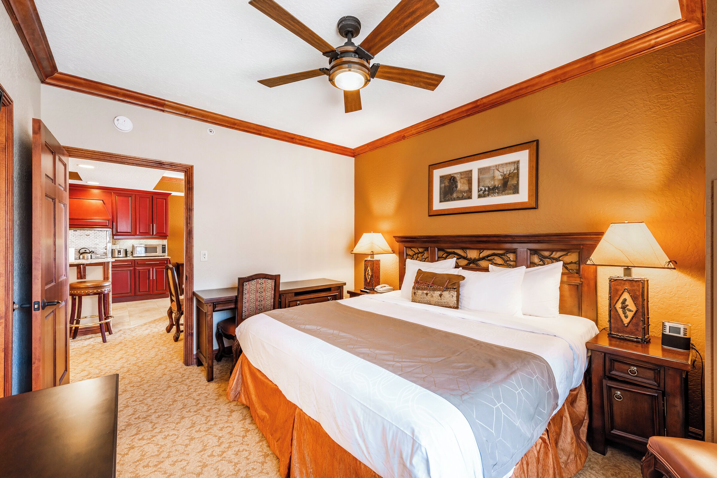 A hotel room with a large bed, wooden headboard, two bedside tables with lamps, a ceiling fan, and a view of the kitchen through an open door.