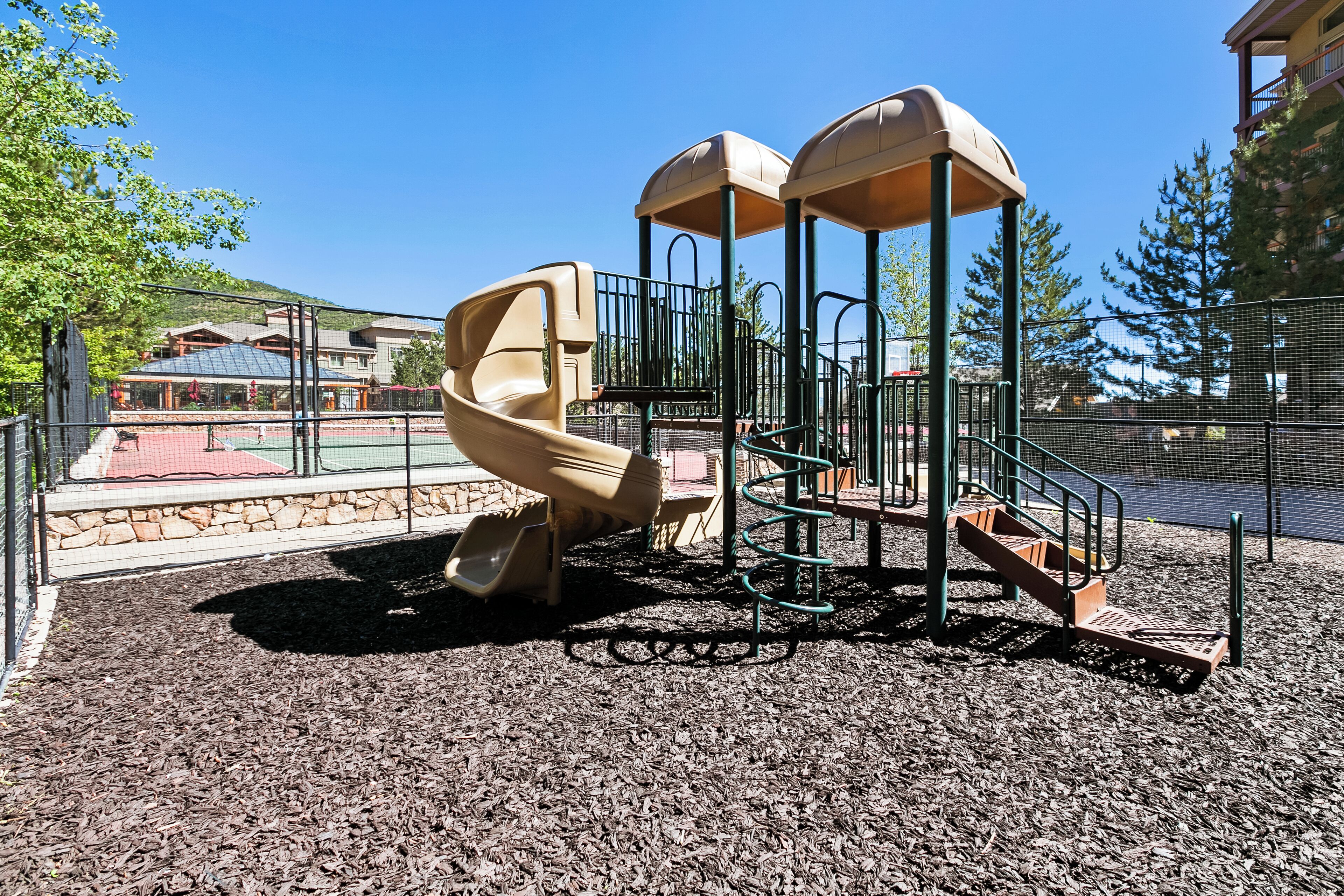 A playground with a slide and climbing structures, surrounded by a fence and mulch ground cover.