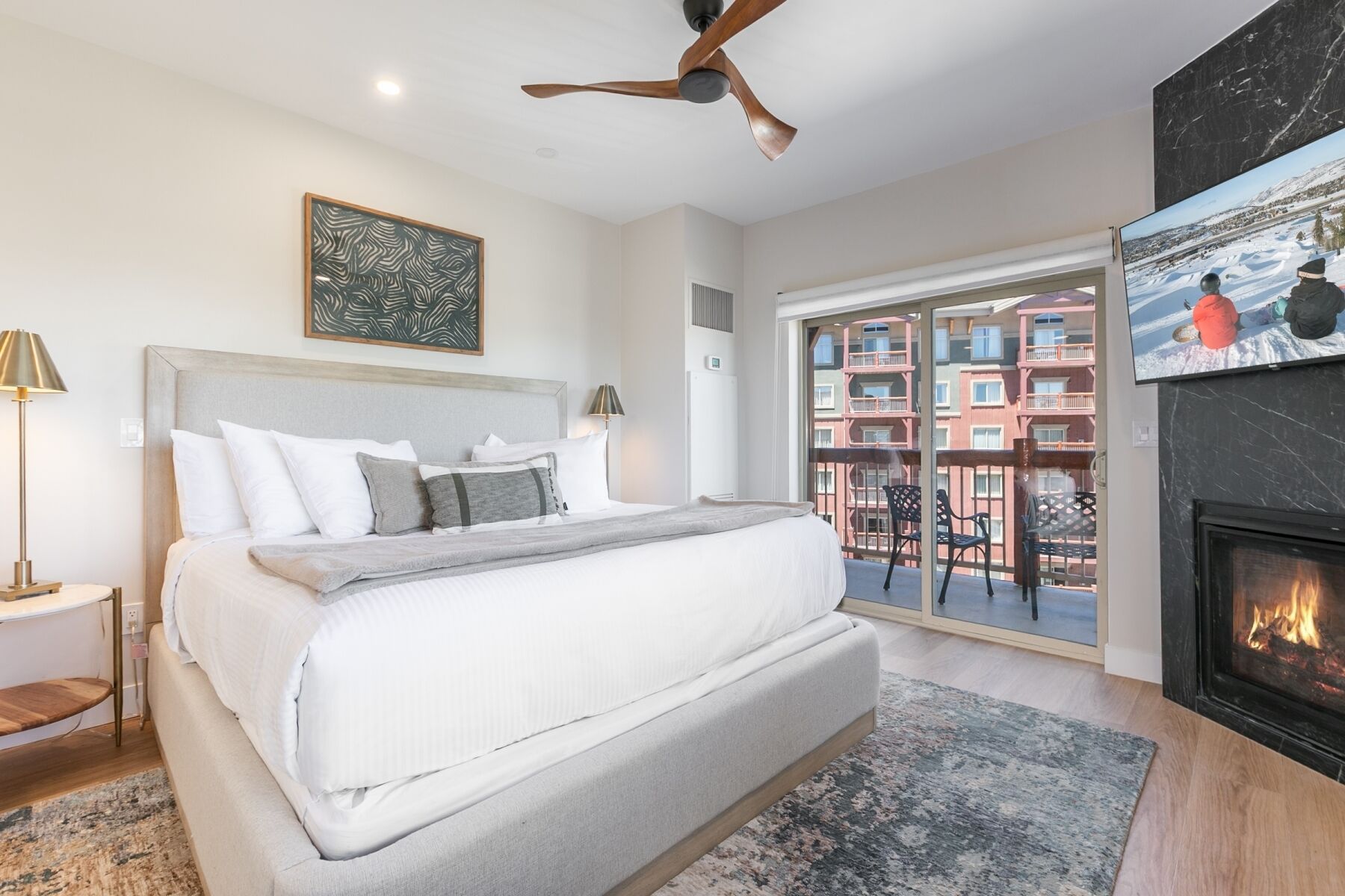 A bedroom with a bed, a ceiling fan, a fireplace, a TV, and a balcony with a view of buildings.