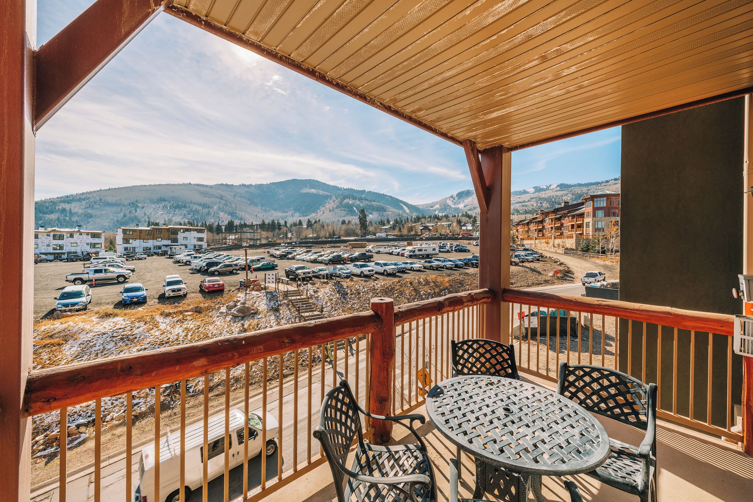 A balcony with a table and chairs, overlooking a parking lot and buildings in a mountainous area.