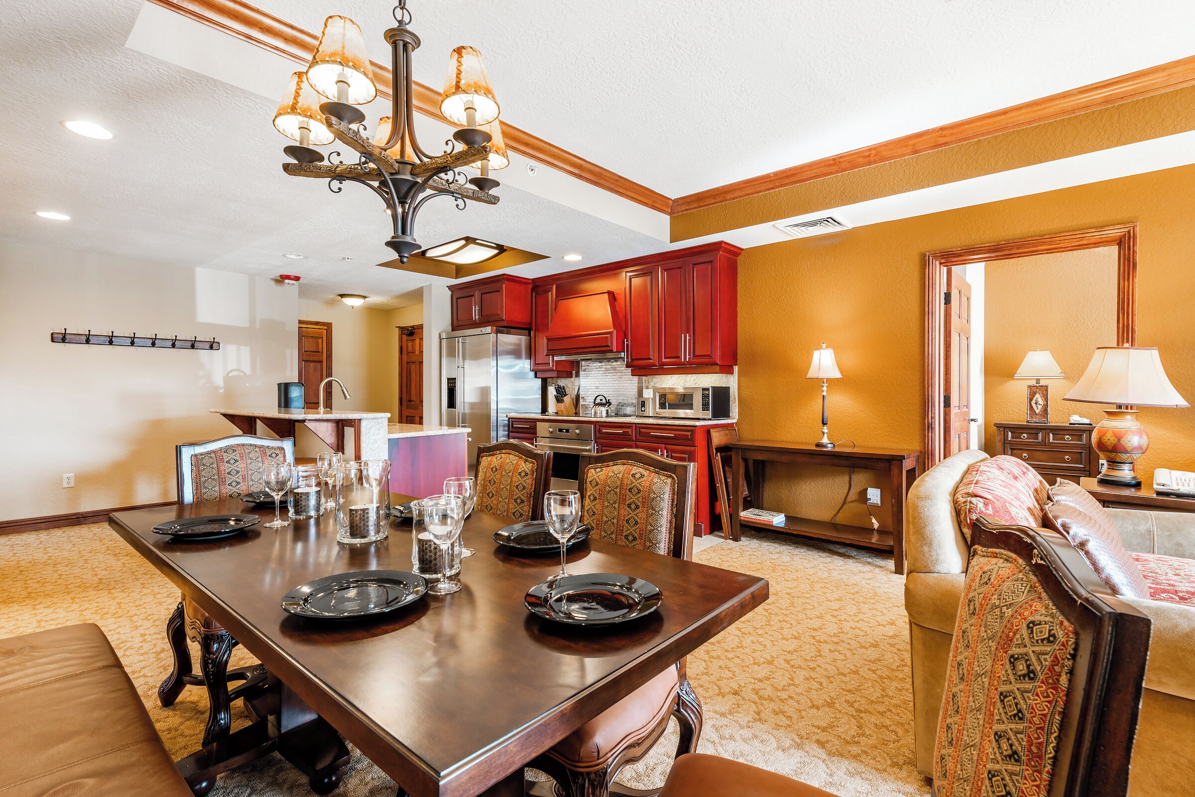 A dining area with a large wooden table, chairs, and a chandelier. There is a kitchen in the background with red cabinets and a refrigerator.