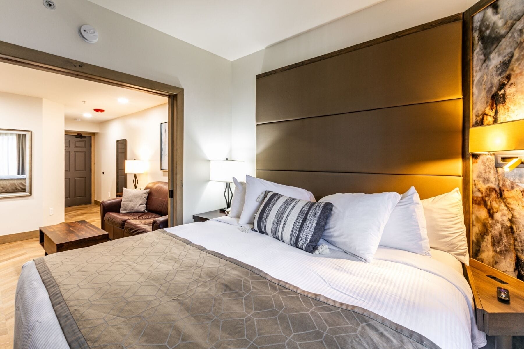 A hotel room with a large bed, a wooden headboard, a nightstand with a lamp, a leather armchair, and a door leading to another room.