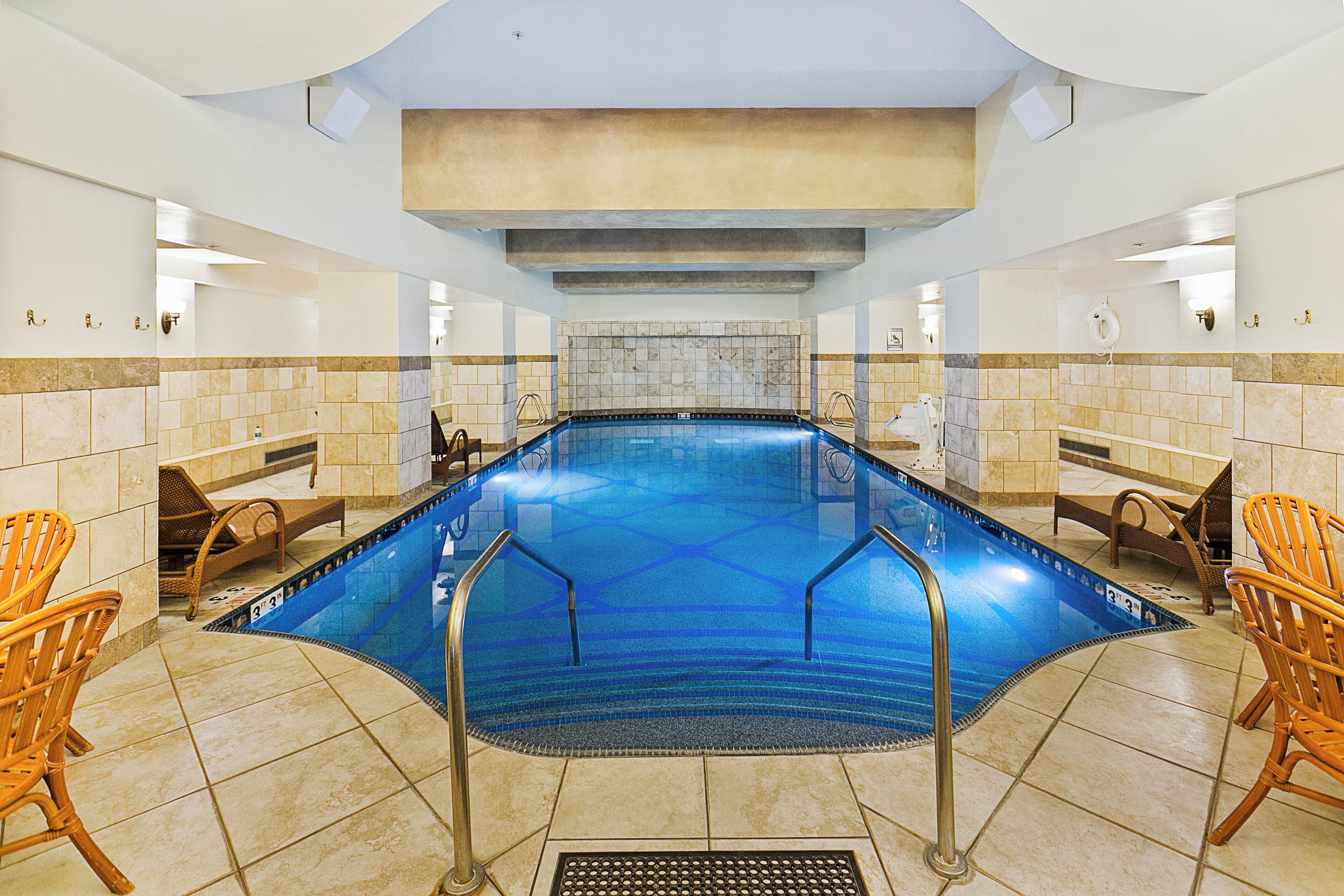 Une grande piscine intérieure à l’eau d’un bleu limpide, entourée de murs recouverts de carreaux beiges et surmontée d’un plafond haut.