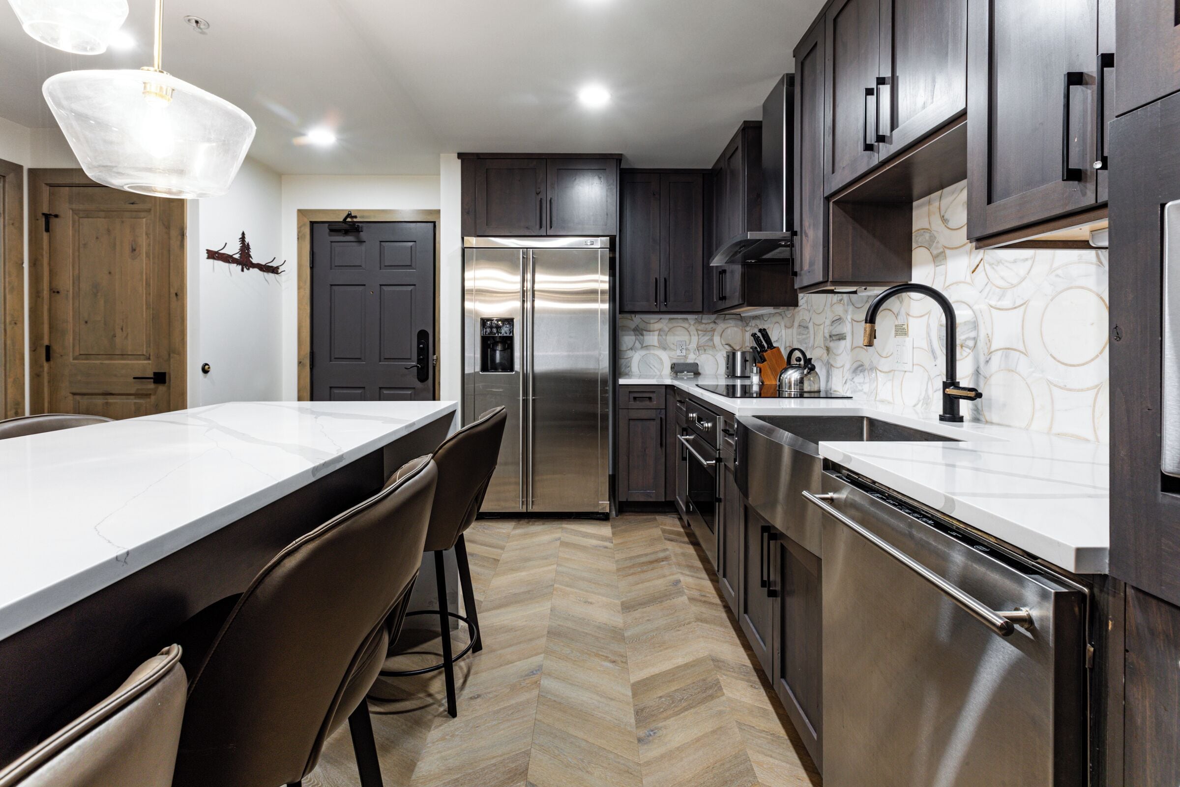 A modern kitchen with a large island, dark wooden cabinets, stainless steel appliances, and a herringbone floor pattern.