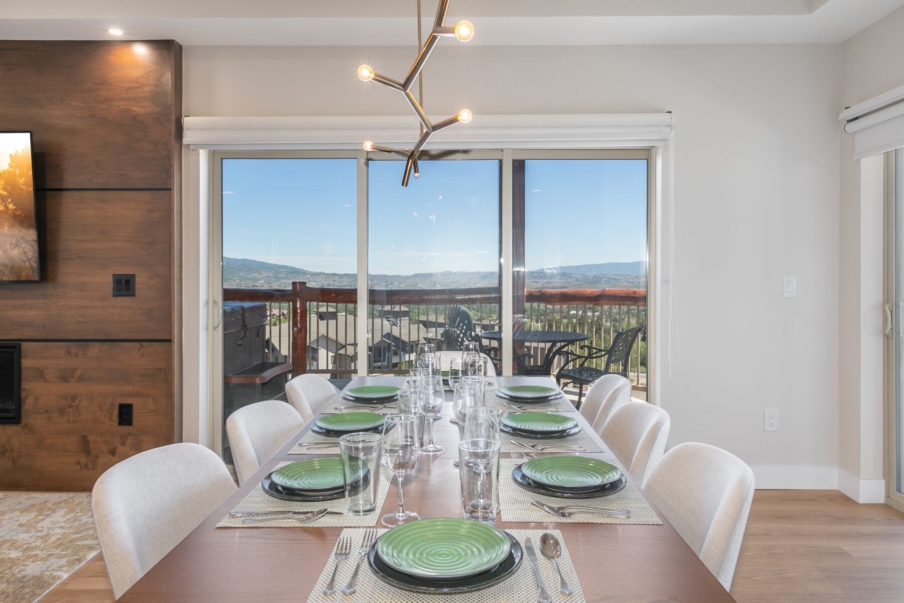 Un comedor con una larga mesa dispuesta para seis personas, ventanas amplias con vistas a un paisaje urbano y una lámpara de araña moderna.