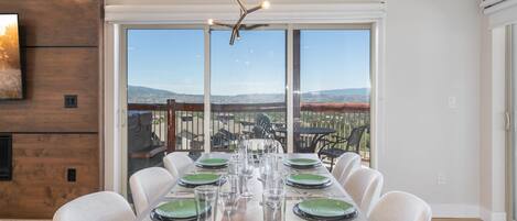 Un comedor con una larga mesa dispuesta para seis personas, ventanas amplias con vistas a un paisaje urbano y una lámpara de araña moderna.