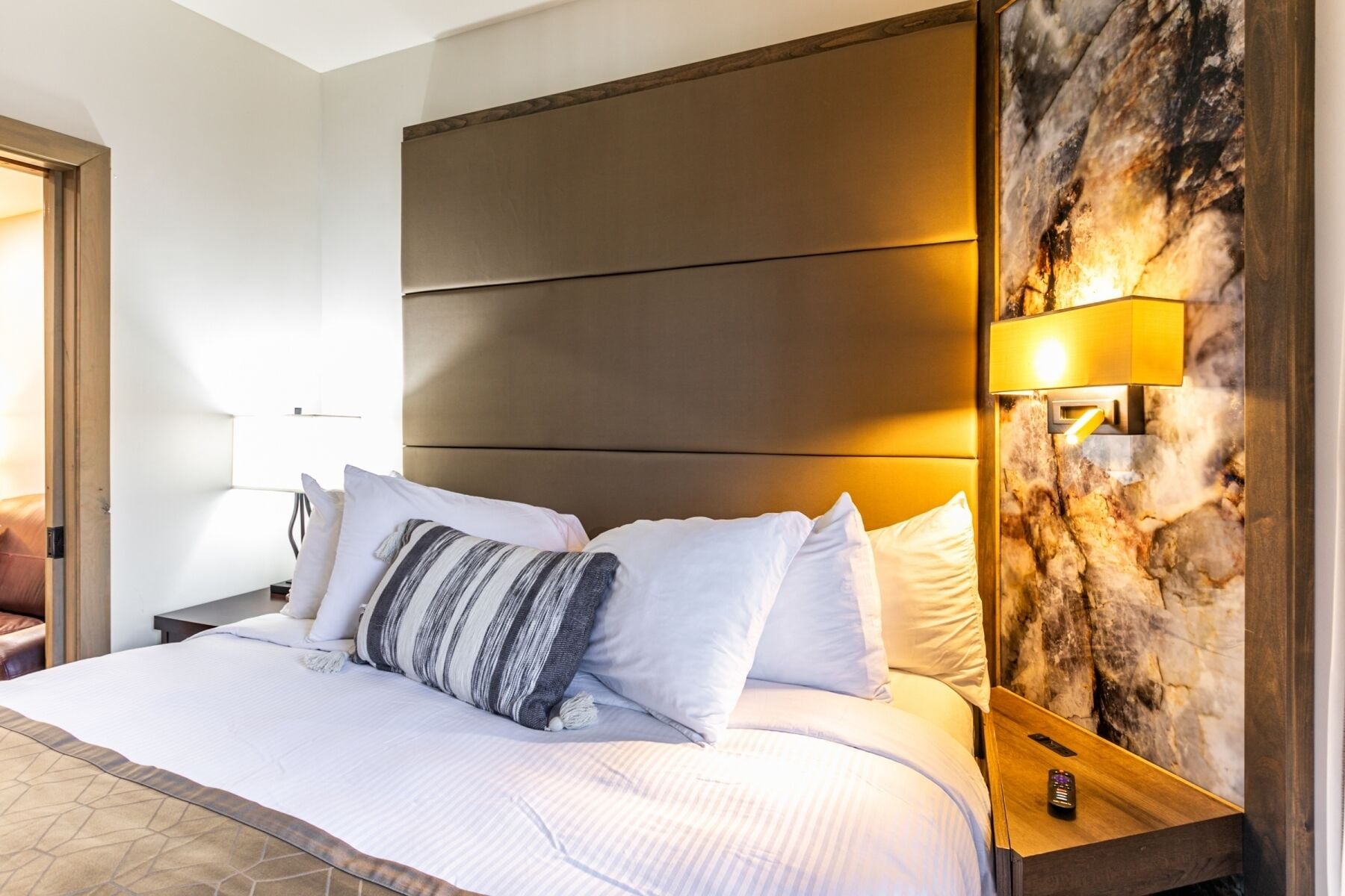 A hotel room with a large bed, a headboard with horizontal panels, a marble accent wall, and a bedside table with a lamp.